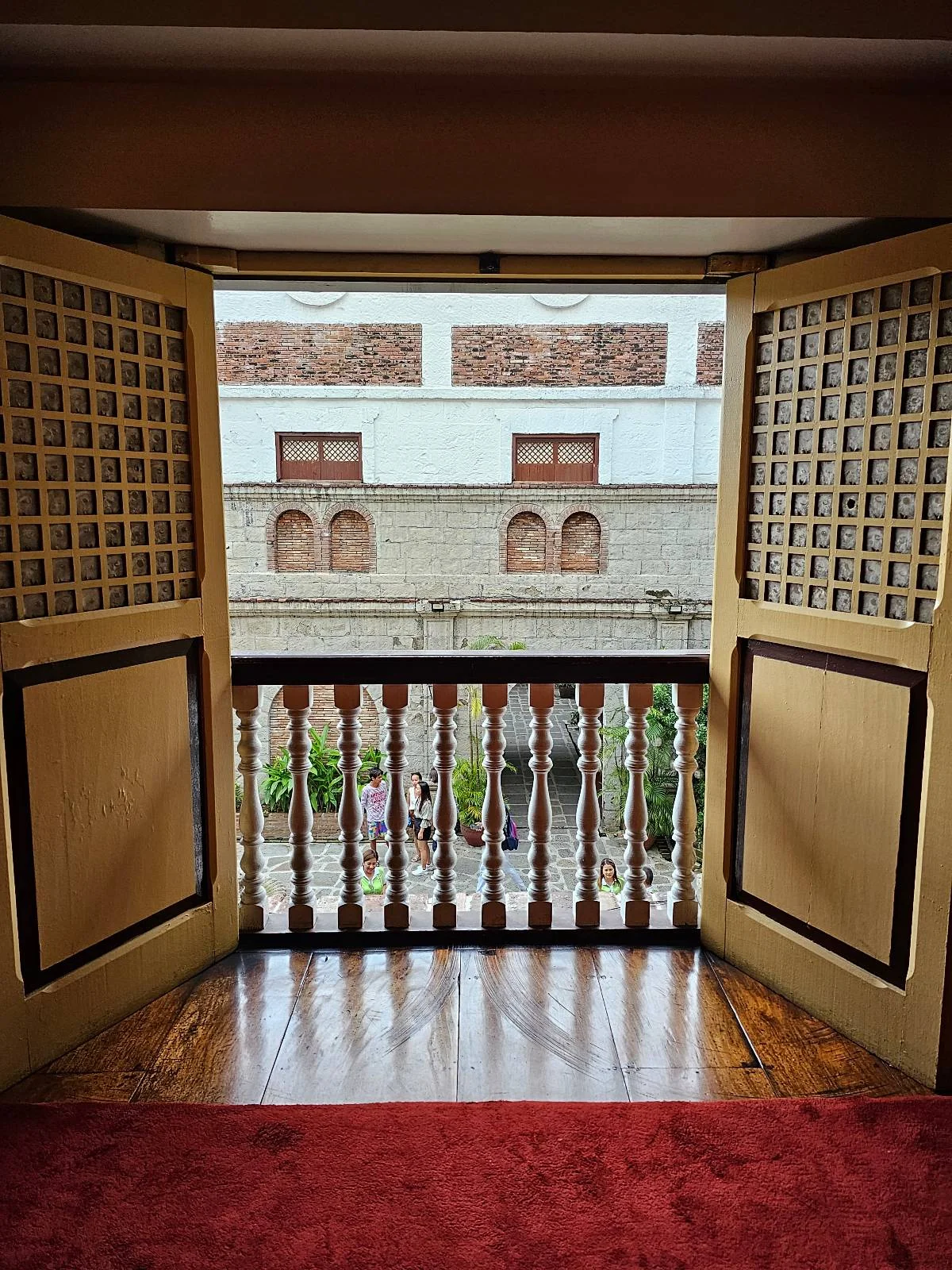 A view from an open wooden balcony door overlooks a street with brick and stone buildings. The door features decorative lattice panels, and the floor inside has a red carpet.