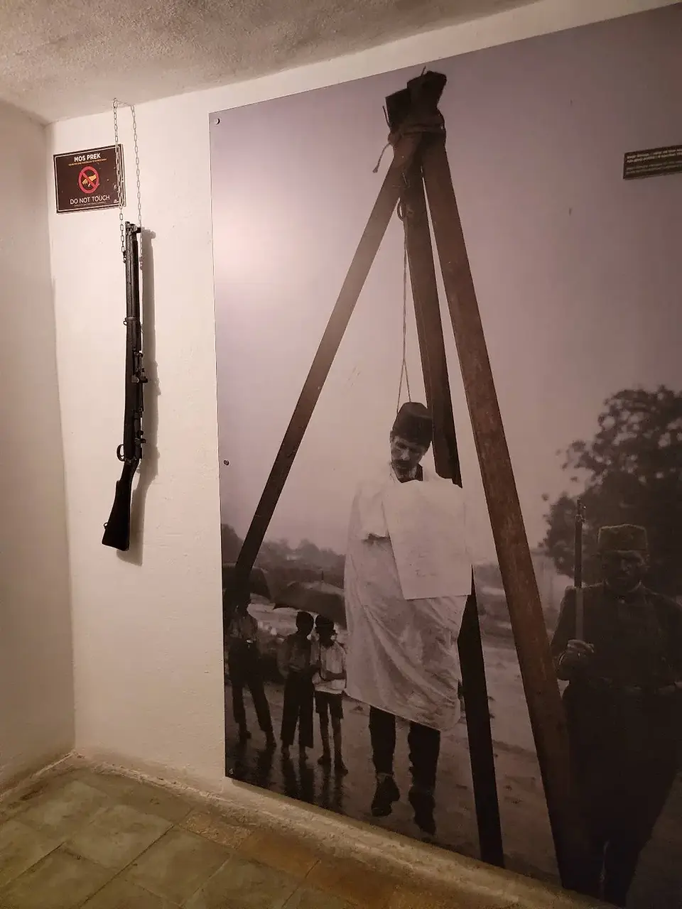 A sepia-toned image on the wall depicts a historical scene with a gallows and a figure in white. In the foreground, there is a rope and plaque display. The room has a minimalist, somber ambiance.