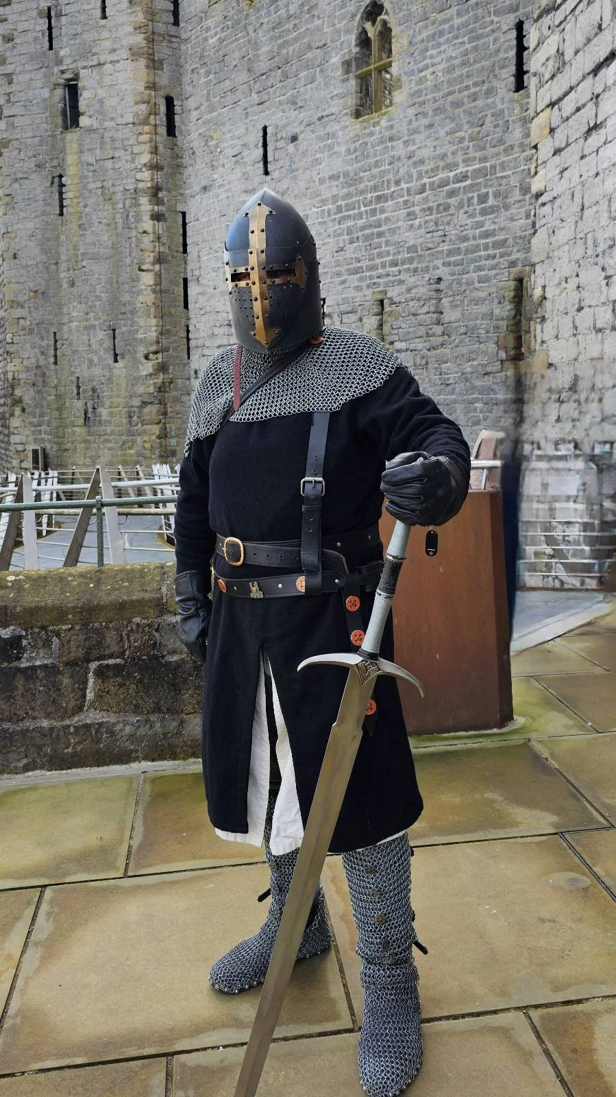 A person dressed as a medieval knight stands on stone tiles, wearing a helmet, black tunic, gray armor, and holding a large sword in one hand, with a stone wall in the background.