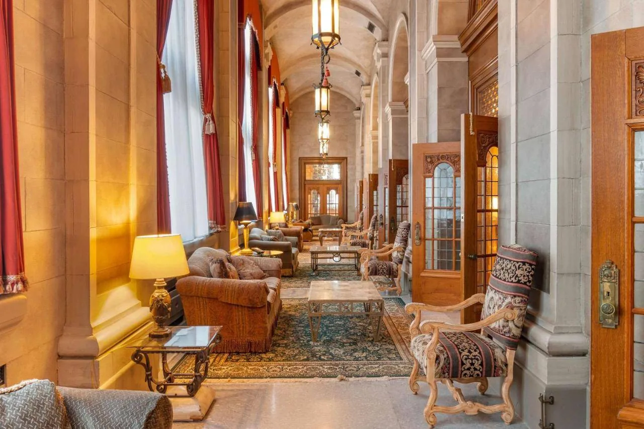 A luxurious hallway with arched ceilings and large windows draped with red curtains. The space is furnished with plush sofas, ornate wooden chairs, and tables. Warm lighting from lamps and chandeliers creates an inviting atmosphere.