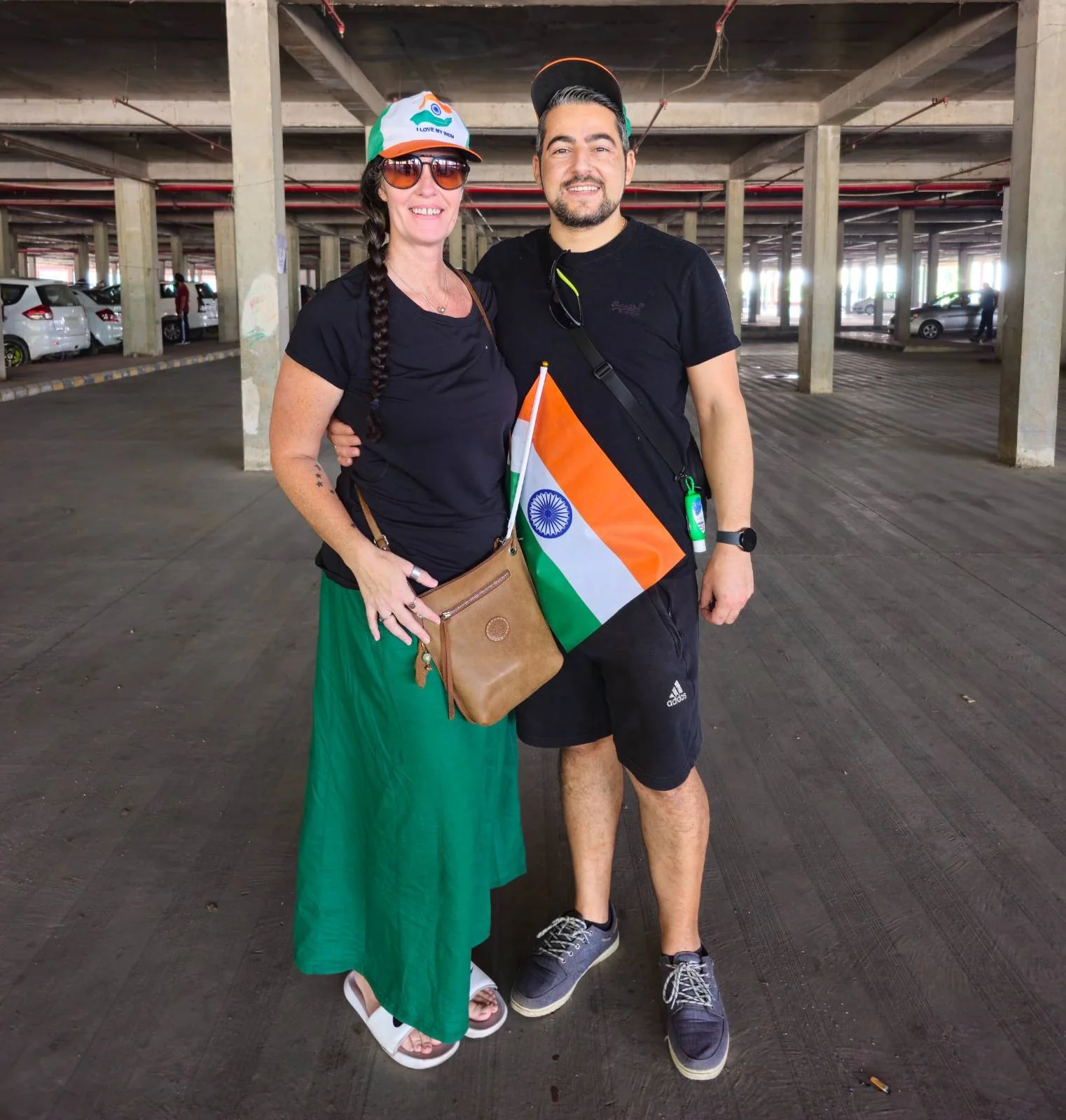 Two people stand together in a parking garage, both wearing casual black tops. One wears a green skirt and a hat, the other wears shorts. They hold an Indian flag and both smile at the camera.