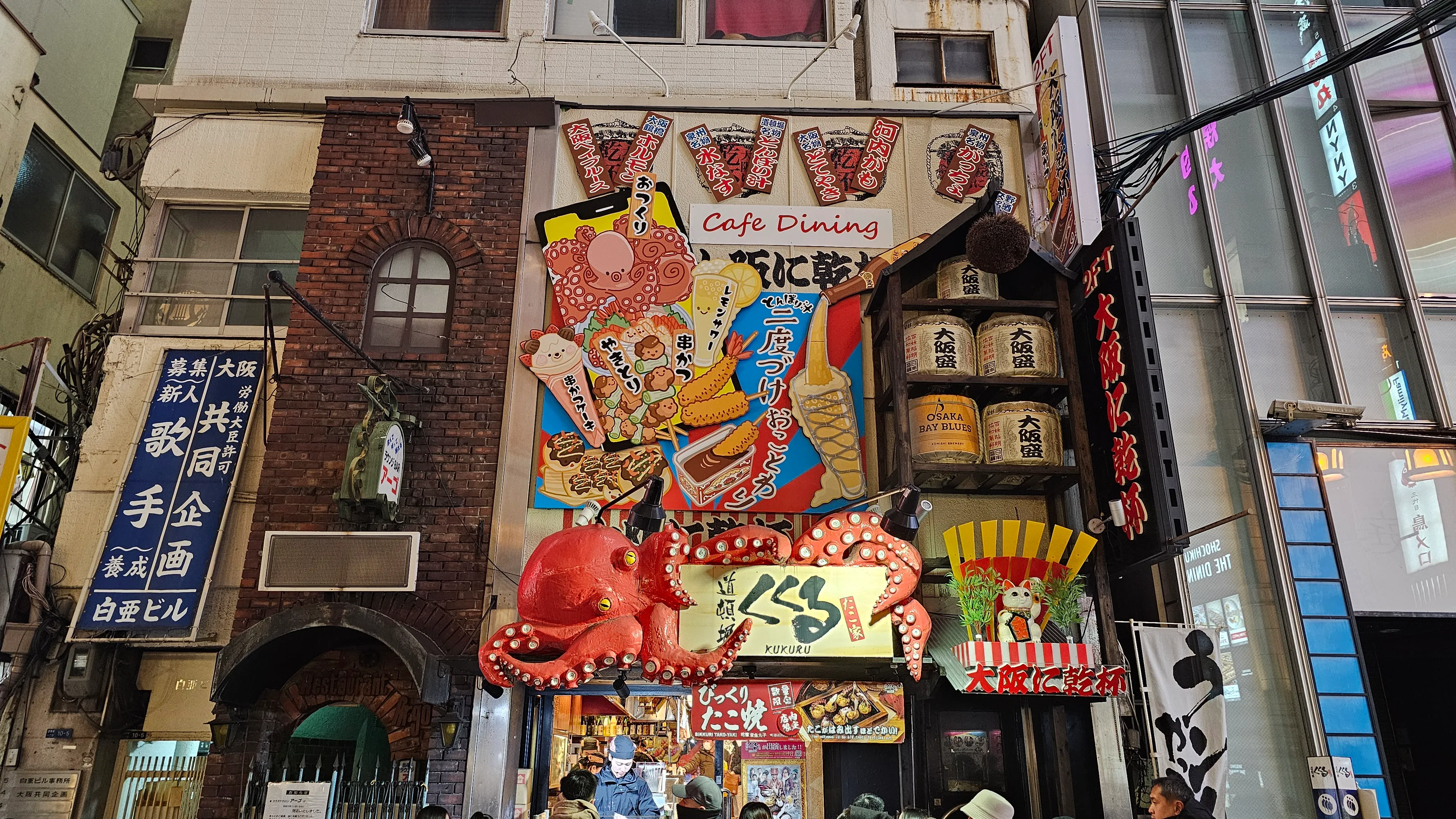 A colorful street scene in Japan featuring bright signs, a large red crab sculpture, and a wall mural of food and drinks above restaurant entrances, with buildings closely packed together.