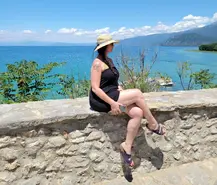 A woman in a black dress and sun hat sits on a stone wall, overlooking the breathtaking views of Ohrid's bright blue sea with mountains beyond. She holds a phone and wears sandals, surrounded by greenery near St. Naum Monastery under a clear blue sky.