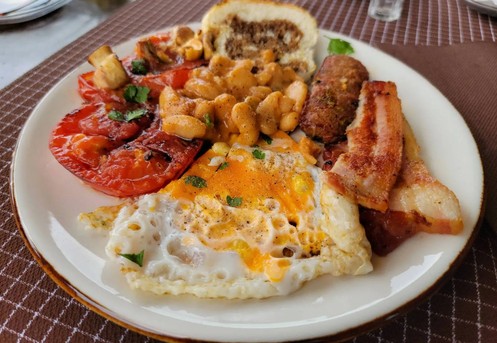 A plate of breakfast food with fried eggs, bacon, cooked tomatoes, beans, sausages, and a slice of stuffed bread. The dish is garnished with herbs and served on a white plate with a brown placemat underneath.