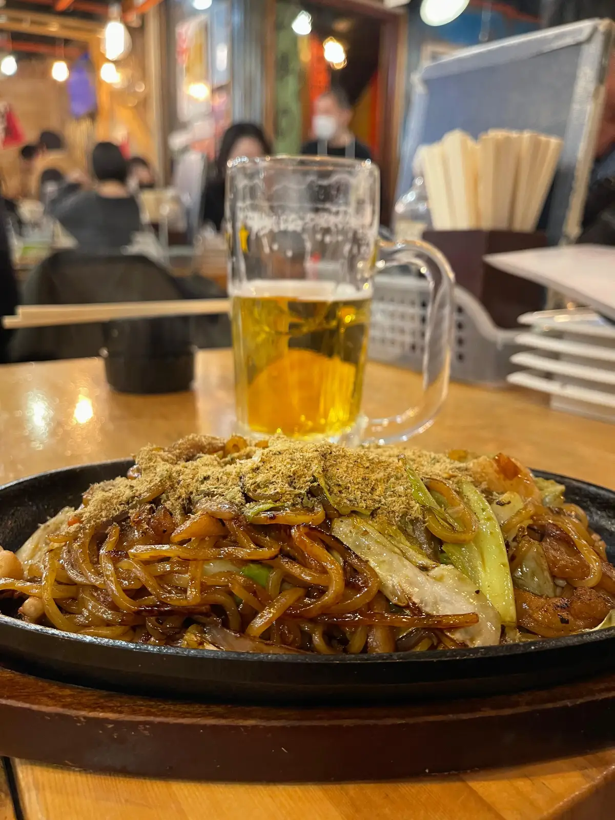A plate of stir-fried noodles topped with vegetables sits on a wooden table, with a pint of beer in a glass behind it; chopsticks and a busy restaurant are visible in the background.