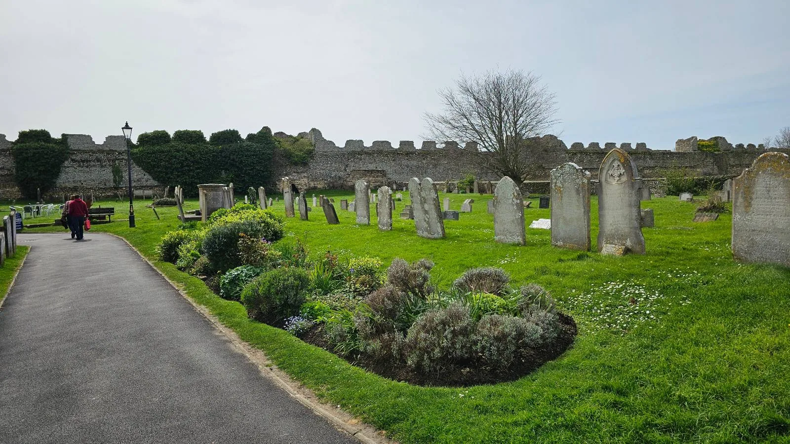 A person walks along a path beside an old cemetery with weathered gravestones and a stone wall, surrounded by lush greenery on a cloudy day.