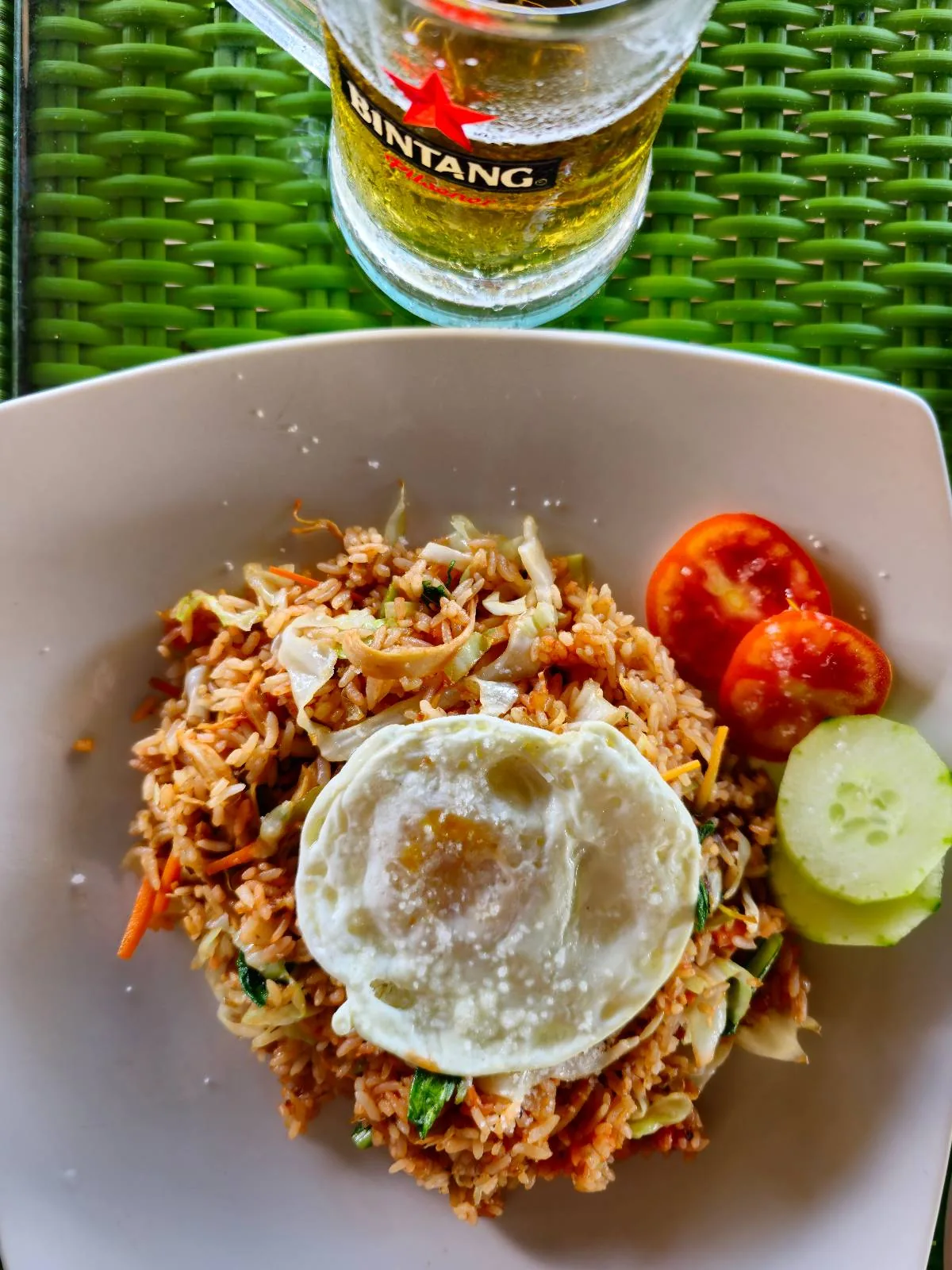 A plate of fried rice topped with a sunny-side-up egg, garnished with sliced tomatoes and cucumbers. A glass of Bintang beer sits in the background on a green woven mat.