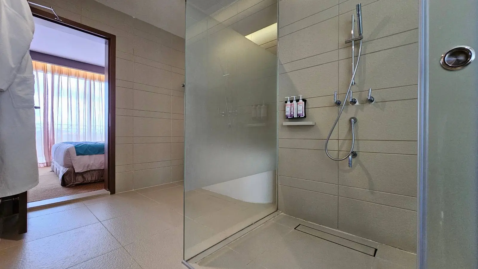 A modern bathroom features a spacious glass-enclosed shower with sleek fixtures and mounted toiletries. The neutral-toned tiles create a clean look, and a glimpse of a bedroom with a neatly made bed is visible through the open door.