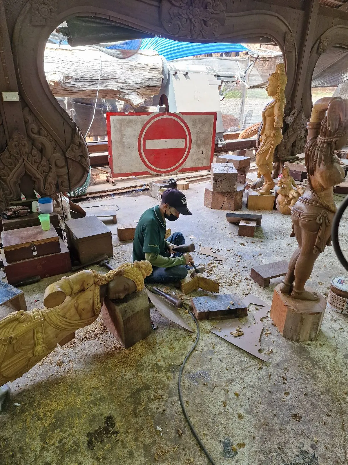 A person is carving wood in a workshop filled with wooden sculptures and carvings. The floor is covered with wood shavings, and a "no entry" sign is visible in the background. A partially carved statue is prominently displayed.