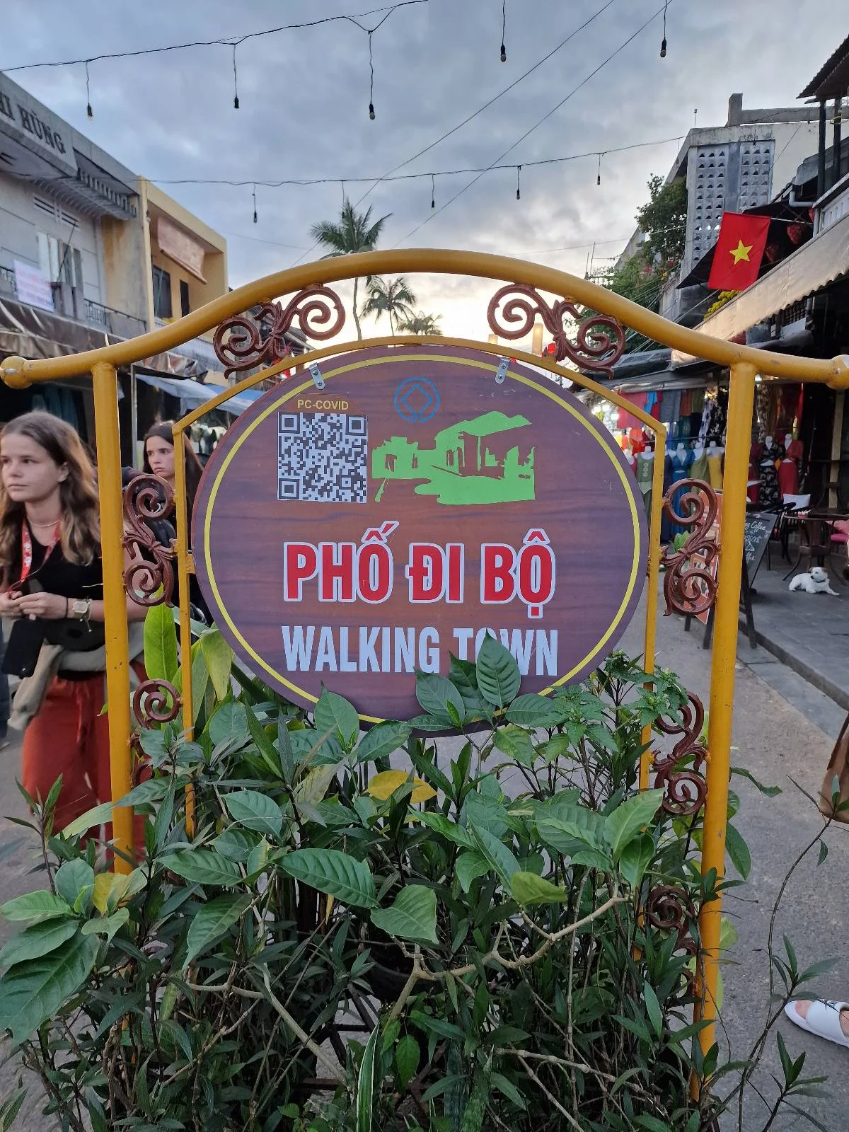 A round sign reads "Phố Đi Bộ Walking Town" in both Vietnamese and English, surrounded by a yellow metal frame with plants in front and street scenes in the background.