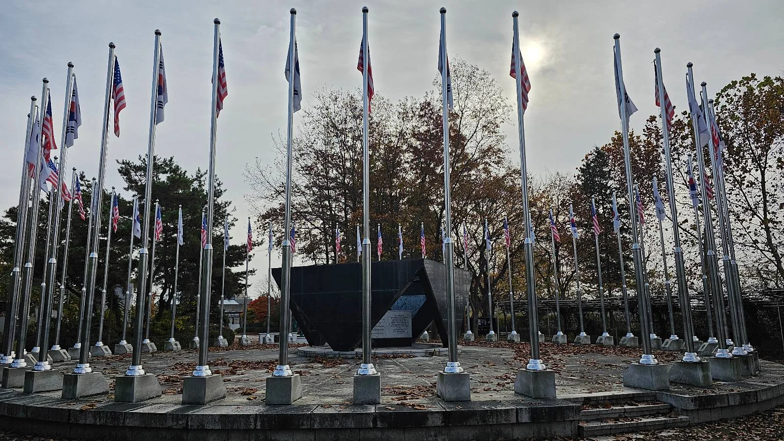 A circular arrangement of flagpoles displaying various flags surrounds a dark stone monument. Autumn trees with sparse leaves are in the background under a cloudy sky.