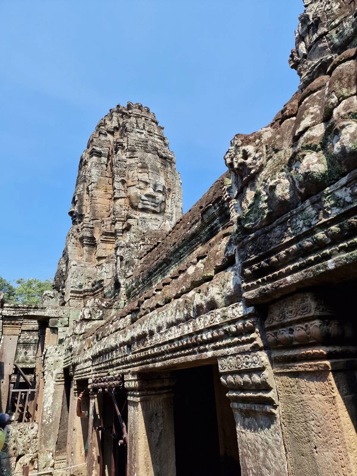 Ancient stone temple ruins with intricate carvings and a tall central tower, set against a clear blue sky at Bayon Temple, Cambodia.