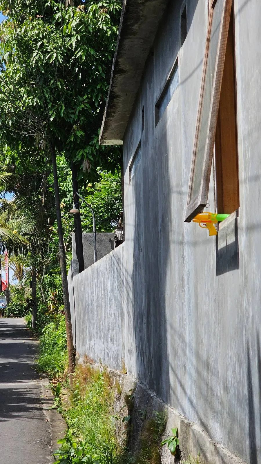 A narrow street with a tall concrete wall on the right. A window on the wall has an open wooden shutter. Green trees and plants line the street, and a colorful street banner is visible in the distance on the left.
