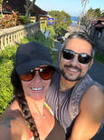 A woman in a black cap and sunglasses smiles alongside a man in black sunglasses on a sunny street. As digital nomads, they embrace slow travel, enjoying the greenery and stone wall with a scenic view of the ocean and buildings in the background.