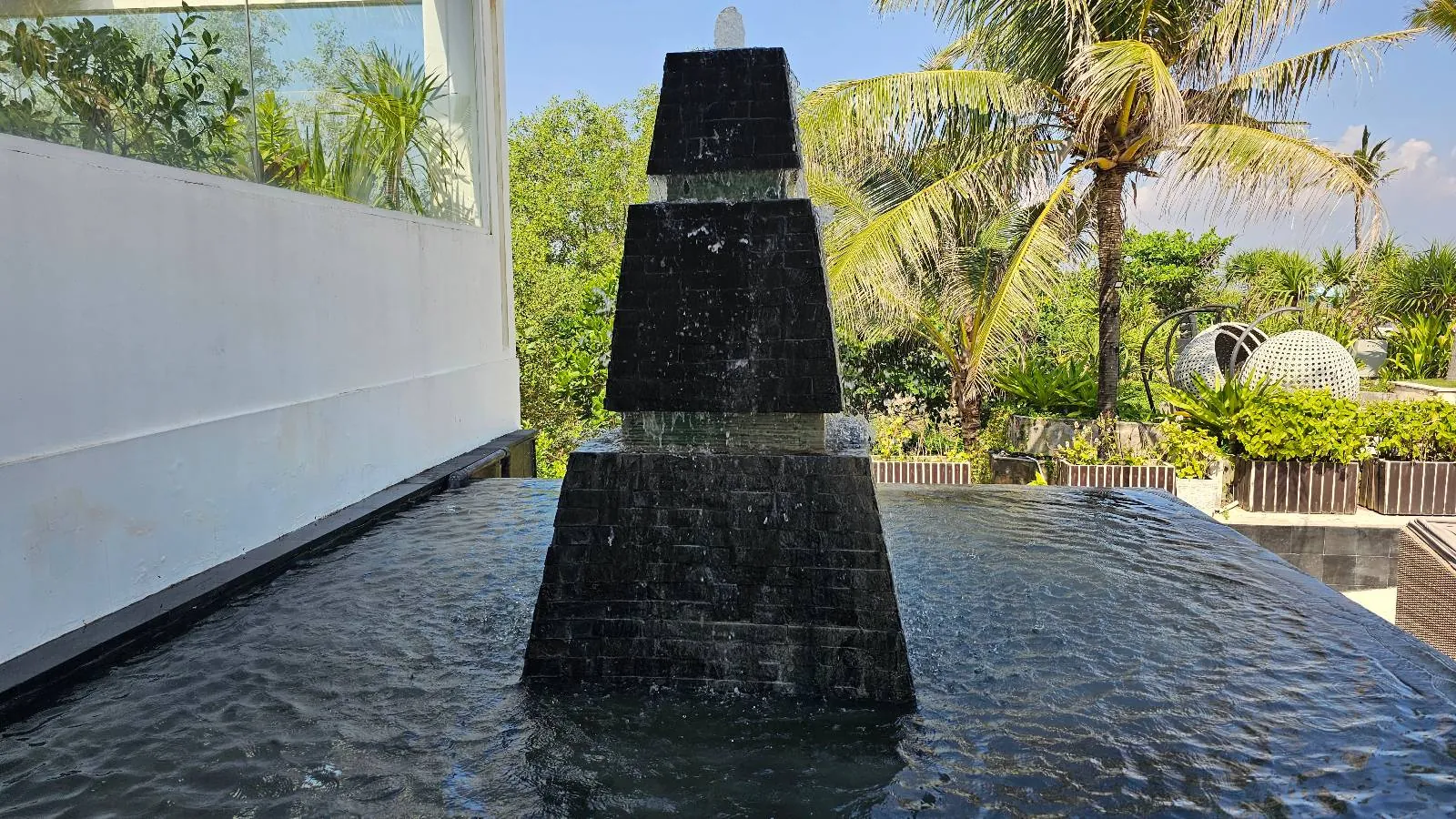 A black, tiered fountain stands on a rippling water surface. It's located on a rooftop or balcony with green trees and palm leaves in the background under a blue sky.