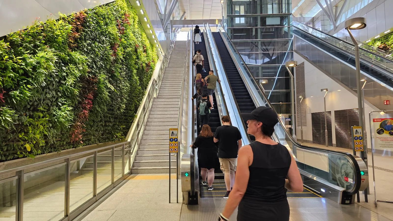 People using escalators in a modern building with a green vertical garden on one side. A woman in a black outfit stands in the foreground. Bright lighting and architectural features create a spacious atmosphere.
