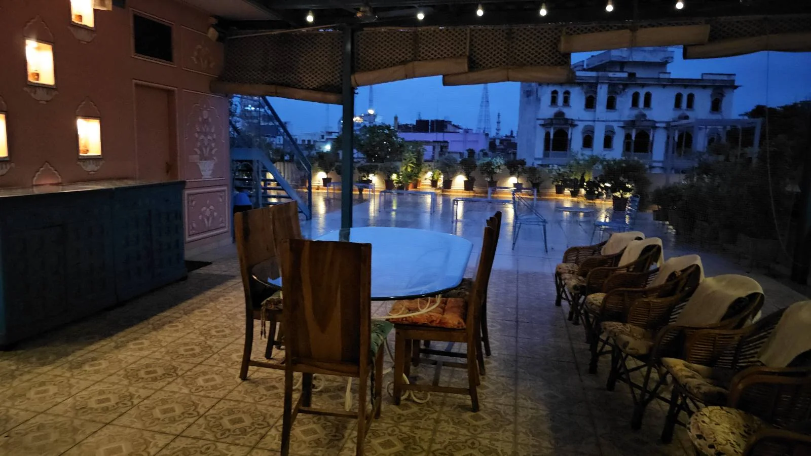 Outdoor rooftop area at dusk with a round table and wooden chairs, string lights overhead, potted plants, and buildings visible in the background under a twilight sky.