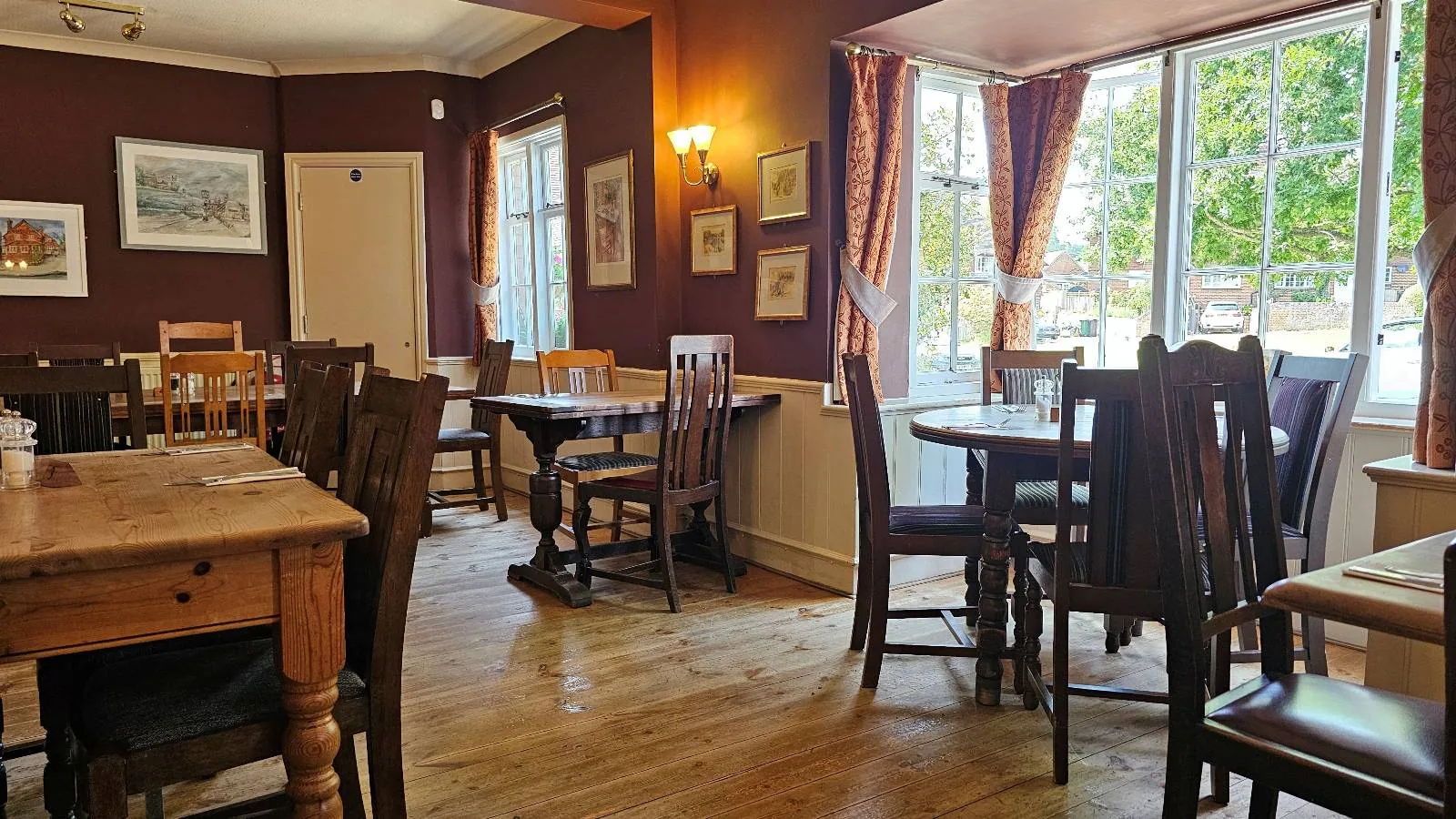 A cozy, sunlit restaurant dining area with wooden tables and chairs, framed pictures on maroon walls, large windows with curtains, and a view of greenery outside.