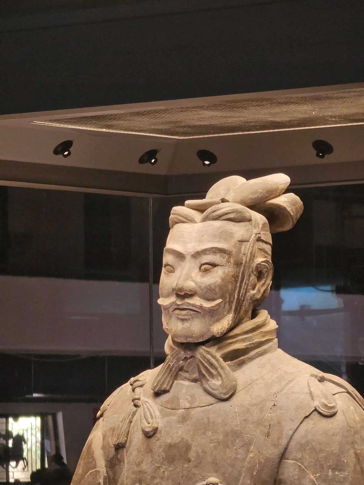 A close-up view of a Terracotta Warrior statue, showing detailed facial features and traditional armor, displayed in a museum setting.