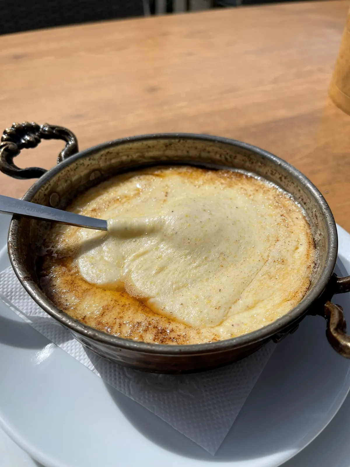 A bowl of melted cheese in a decorative metal dish with handles on a white plate, placed on a wooden table. A spoon is resting in the cheese.