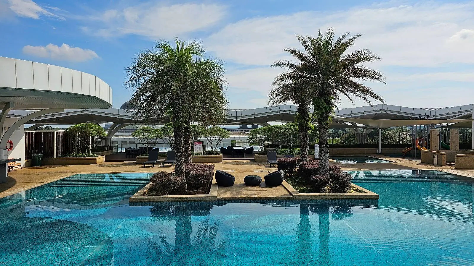 A luxurious rooftop pool features two palm trees on a small island, surrounded by clear blue water. The setting includes modern architecture and lounge chairs, with a partly cloudy sky overhead.