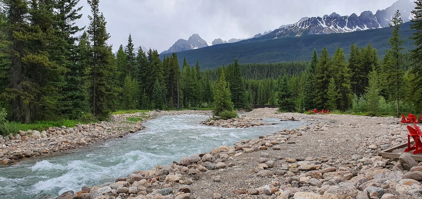 A rocky riverbank with a flowing river, surrounded by tall, green pine trees and distant mountain peaks under a cloudy sky. Several red chairs are scattered along the riverbank.