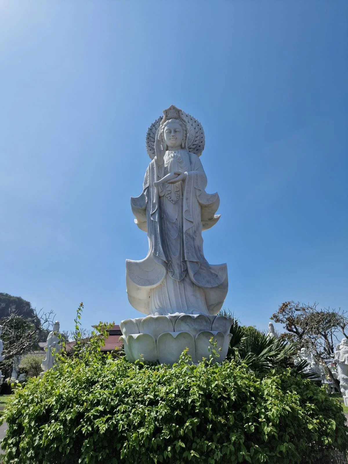A tall, white statue of a robed figure stands on a platform surrounded by greenery under a clear blue sky.