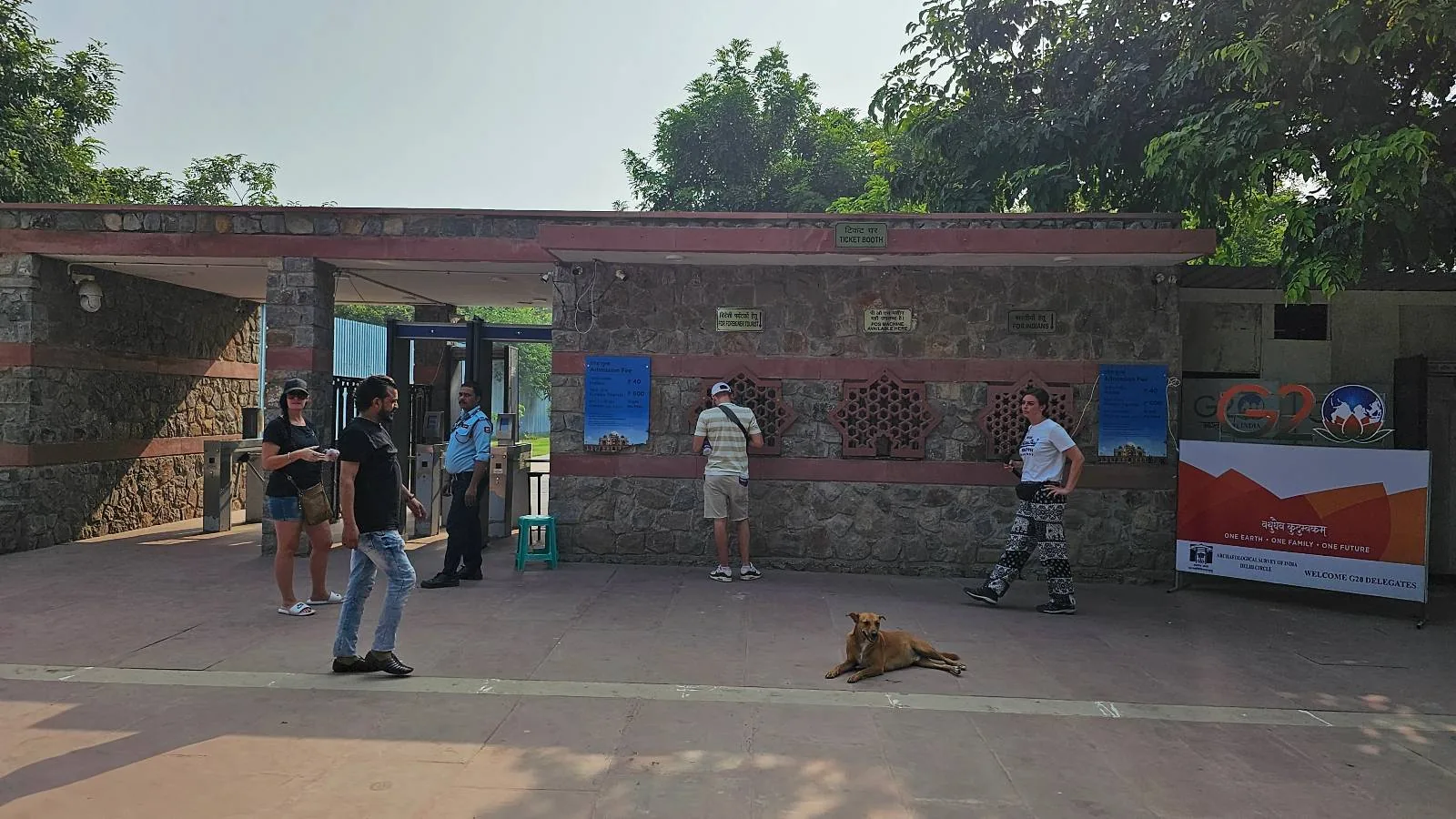 A ticket booth near a gated entrance with "Humayun's Tomb" written on a wall in multiple languages. Two people stand by the booth, and an auto-rickshaw is visible in the background. A dog rests on the ground nearby.