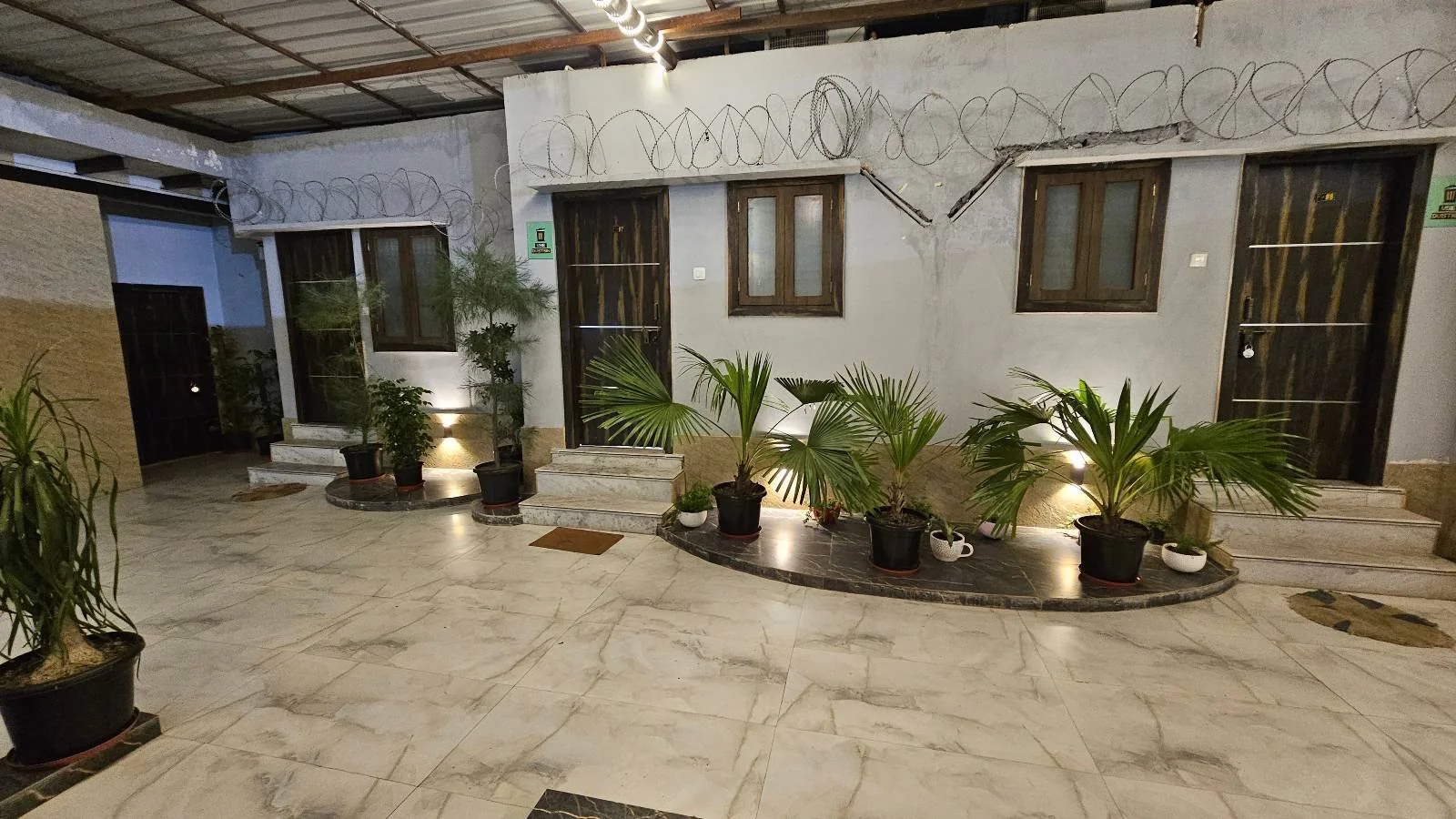 A courtyard with a marble floor features potted plants arranged in a semicircle. Two doors with barred windows are on the back wall, under a corrugated metal roof. The area is well-lit with overhead and ground lighting.
