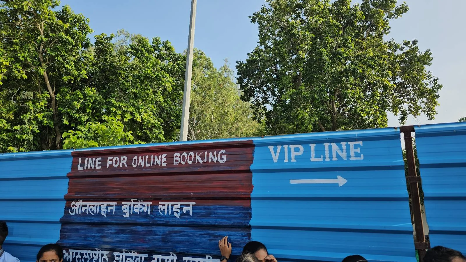 A blue partition outdoors shows two signs: "LINE FOR ONLINE BOOKING" in red and "VIP LINE" in white with an arrow pointing right. People stand in front of the barrier, and green trees are visible in the background.