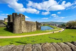 Wide view of Caerphilly Castle in Wales, showing the massive stone walls, surrounding moat, and green parkland under a bright blue sky.