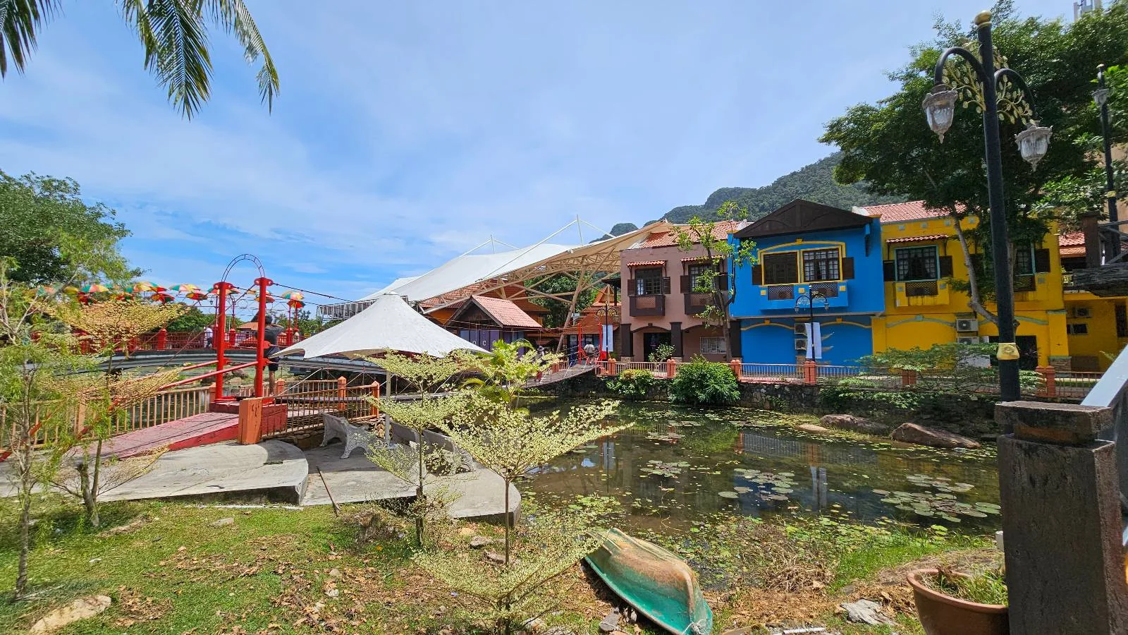 A vibrant setting with colorful buildings in shades of pink, yellow, and blue. A small pond with a few plants sits in the foreground. A white tent, red bridge, and lanterns enhance the lively atmosphere under a clear blue sky.