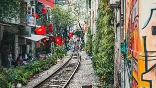 A narrow residential street in Hanoi with a functional railway track running through the center. Buildings on both sides are adorned with Vietnamese flags and lush potted plants, with people sitting at cafes along the tracks.