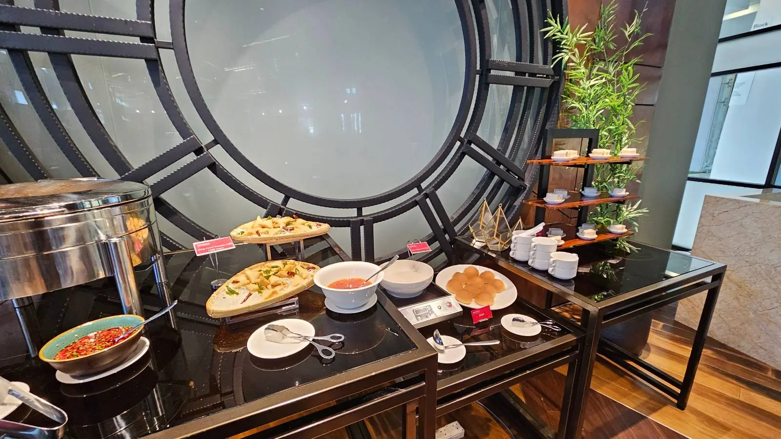A buffet setup features various dishes on black tables, including bowls of soup, plates of pasta, and bread. A metal structure with a circular design is on the wall behind, and a small bamboo plant adds a touch of greenery.