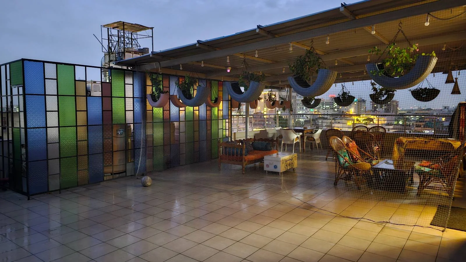 A spacious rooftop terrace at dusk with colorful glass panel dividers, hanging plants, and cozy seating areas under a canopy, overlooking a cityscape.