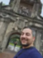 Man smiling in front of historic stone archway with engraved details. Woman in background poses playfully. Overcast sky, lush greenery.