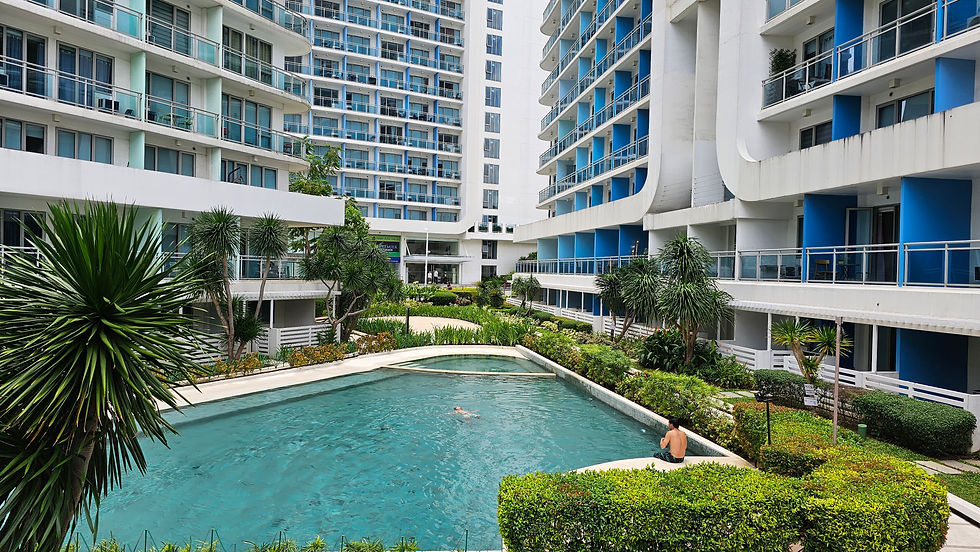 Courtyard pool with a swimmer and man sitting on edge, surrounded by lush greenery and modern white-blue buildings under a cloudy sky.