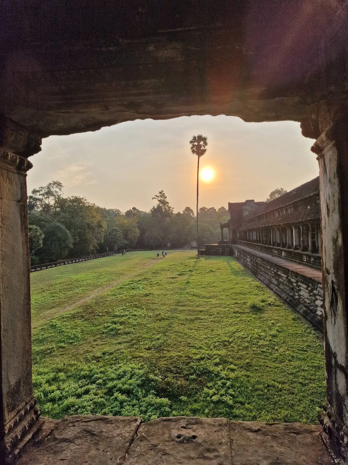 View through a stone archway to a grassy area, with the sun low in the sky near a tall, ancient stone pillar and trees in the distance, suggesting sunrise or sunset at a historic site.