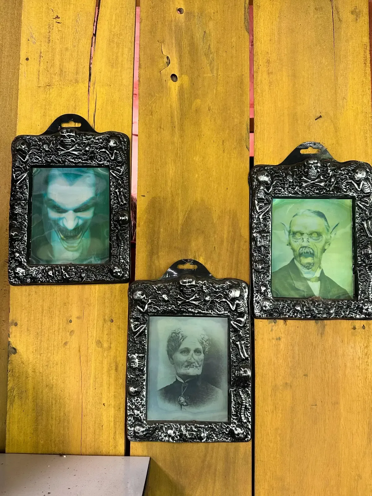 Three ornate, black-framed vintage portraits hang on a yellow wooden wall. The top left portrait appears ghostly or distorted, while the other two show an older man and woman in old-fashioned attire.