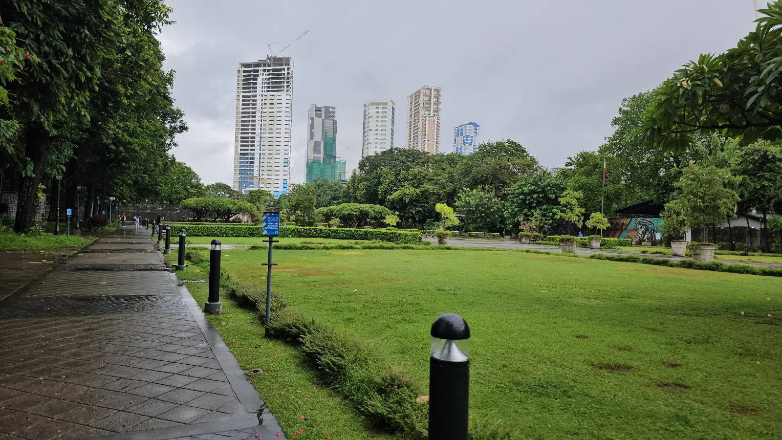 A park with lush green grass and surrounding trees on a cloudy day. A paved walking path runs along the left, with skyscrapers visible in the background. A few scattered park lamps line the path.