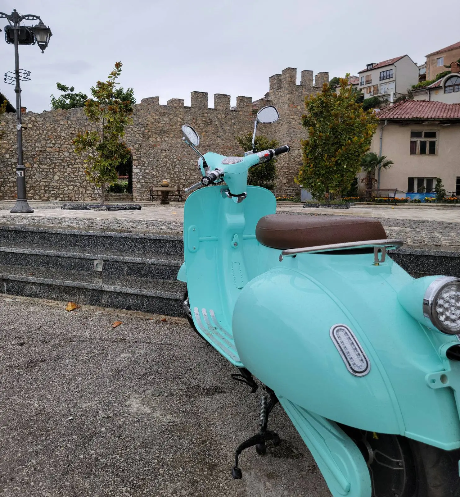 A mint green scooter parked on a cobblestone street. A stone wall with crenellations and a few trees are in the background, along with residential buildings. The sky is overcast.