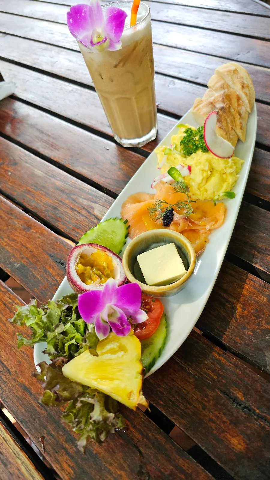 A long plate with scrambled eggs, smoked salmon, fruit slices, salad greens, and a flower garnish. A glass of iced coffee sits on the wooden table next to it.