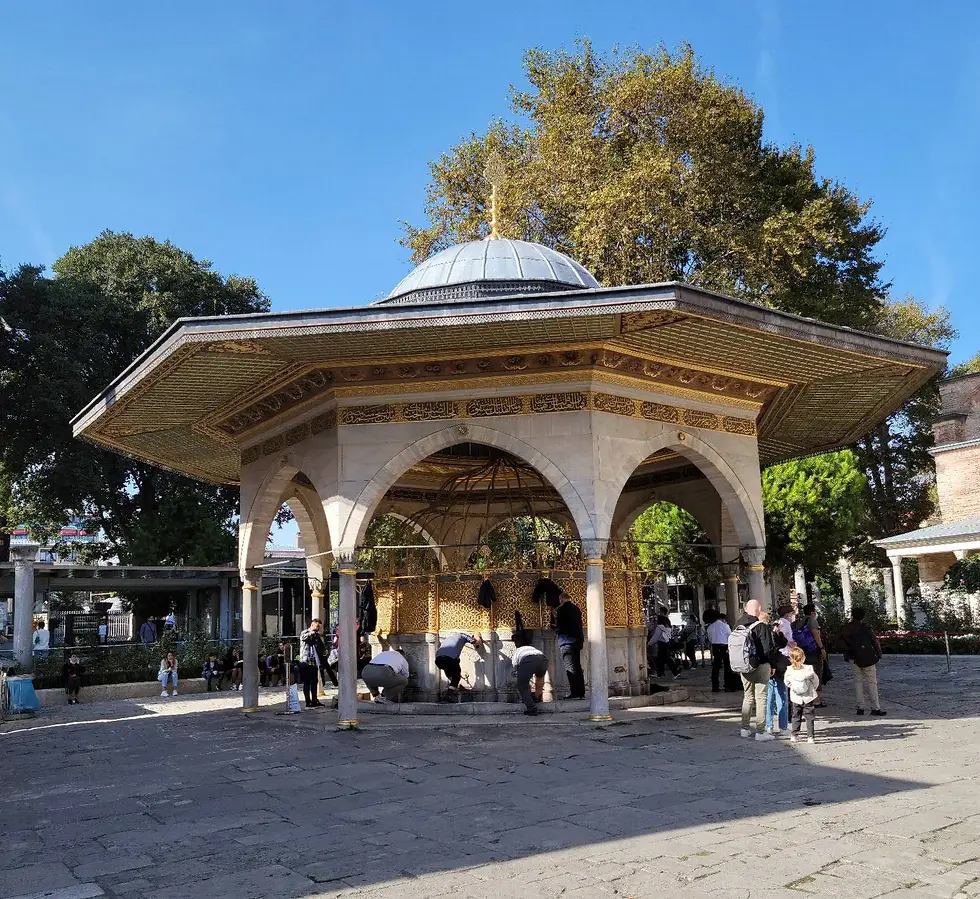 A group of people gather under an open pavilion with arches and a domed roof in a park setting.