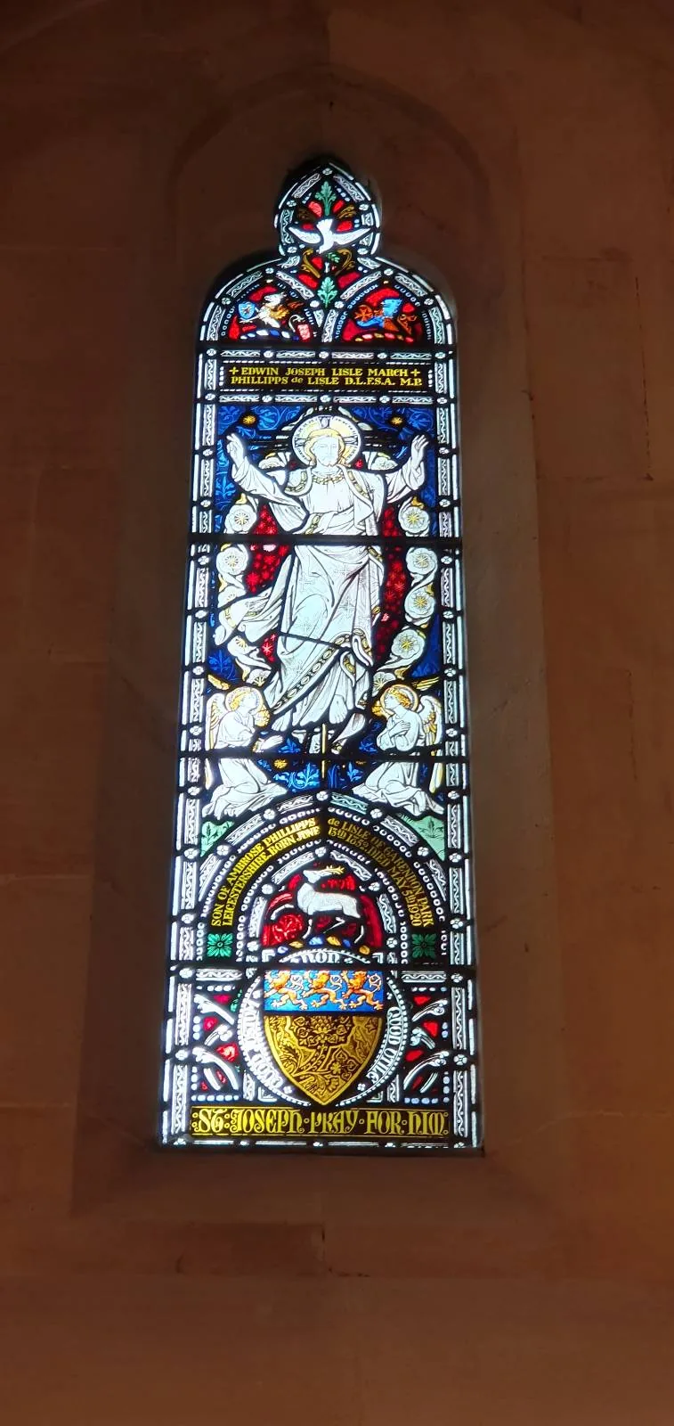 Stained glass window depicting a seated angel in a white robe with red wings, holding a scroll. Below is a colorful shield with symbols. The window features ornate patterns in red, blue, and yellow against a light brown wall.