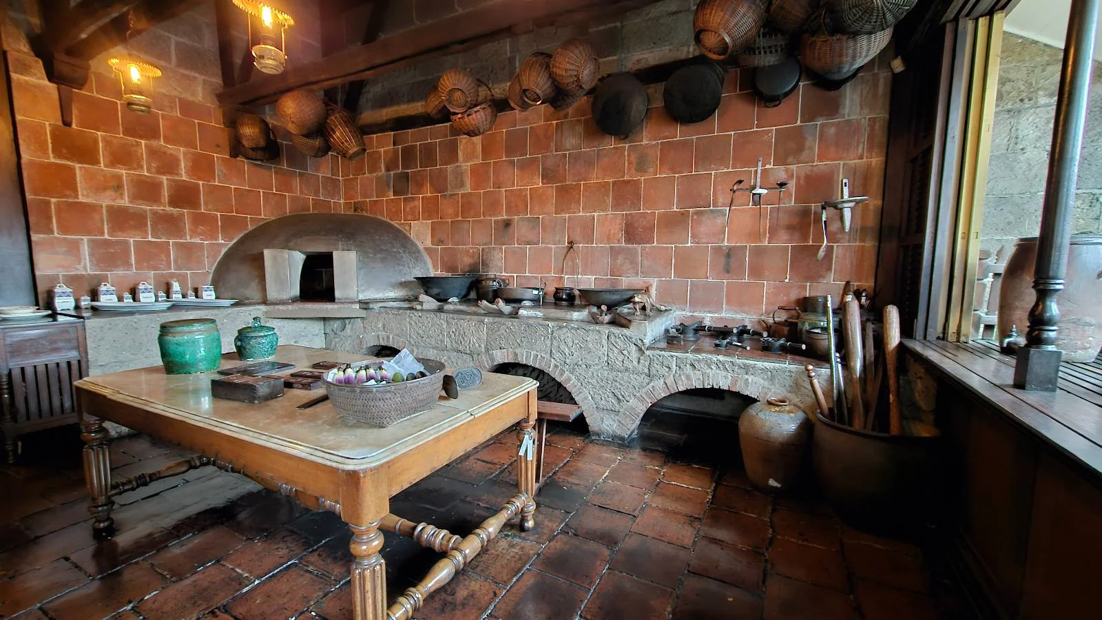 A rustic kitchen with a large brick oven, stone countertops, and a wooden table with kitchenware. Copper pots and utensils hang on the wall, and the floor is tiled. The space looks warm and vintage, with a window on the right letting in natural light.