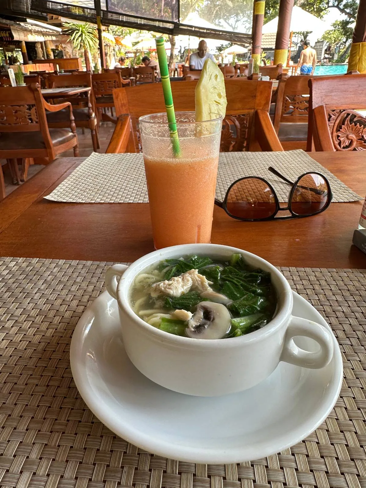 A bowl of soup with greens and mushrooms sits on a white plate on a woven placemat. Behind it, a glass of orange juice with a green straw and a pineapple slice rests on the table. Sunglasses are placed nearby in a restaurant setting.