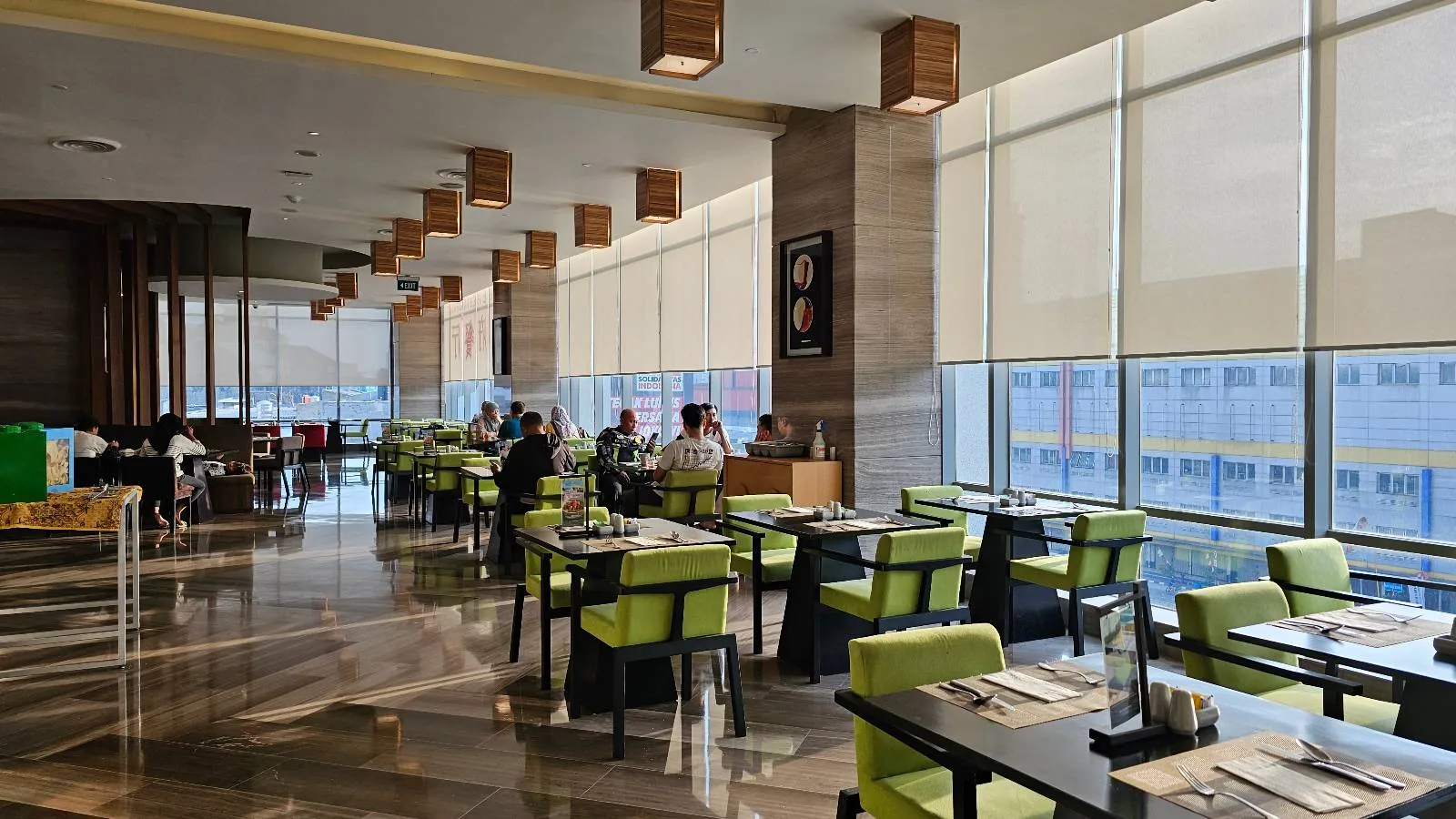 A modern restaurant with floor-to-ceiling windows, green chairs, and neatly set tables. A few people are dining, and sunlight streams in, creating a bright and welcoming atmosphere.