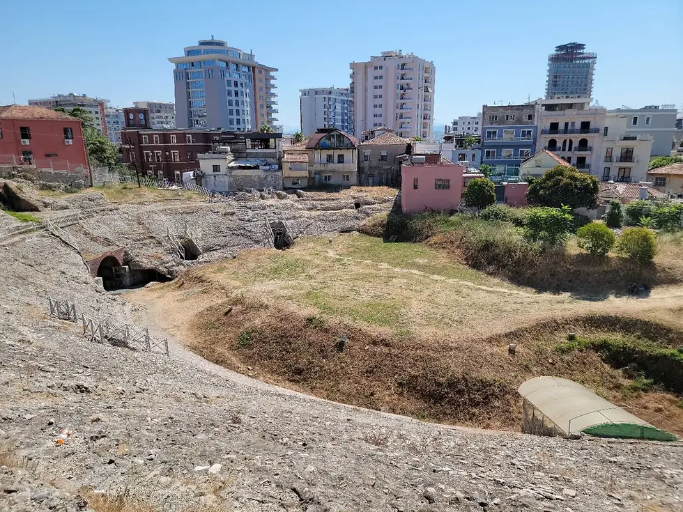 Ancient amphitheater ruins with grass and stone paths, surrounded by modern buildings under a clear blue sky. Mixed historic and urban vibe.