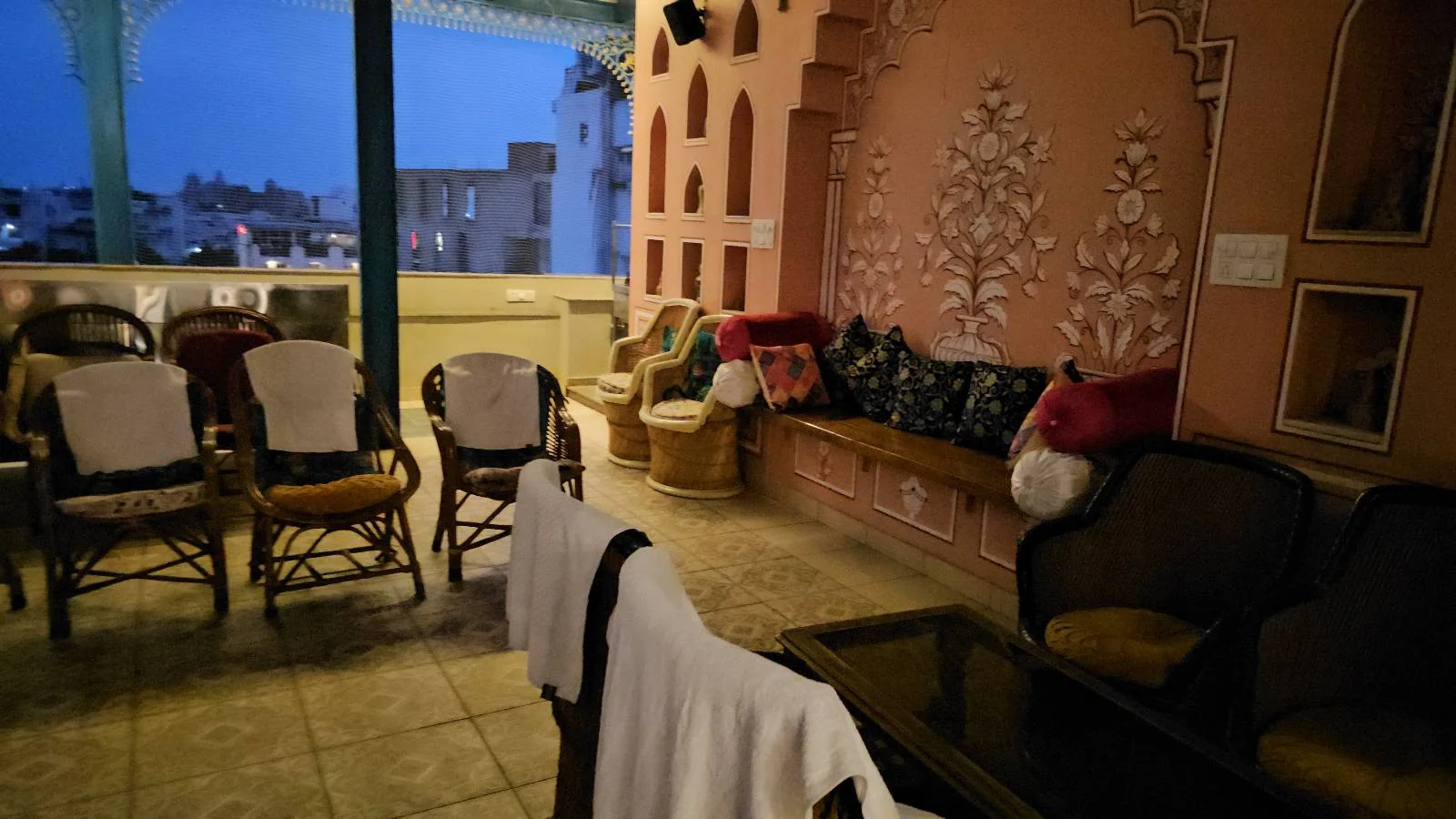 A cozy indoor seating area with wicker chairs and cushioned benches, some draped with white towels, next to a decorative pink wall, and an open balcony overlooking city buildings at dusk.