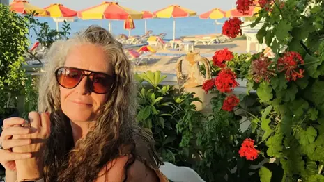 Jenn seated at Mavromatis Taverna in Greece, surrounded by greenery and flowers, with beach umbrellas and sun loungers visible along the shoreline behind her.